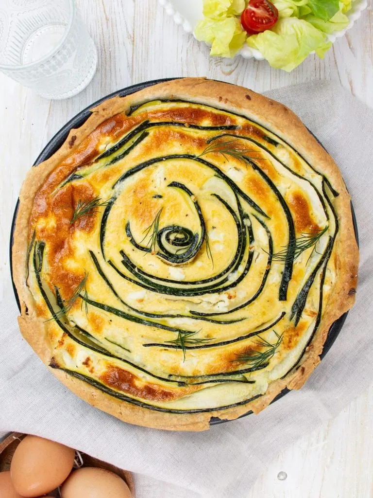 An overhead shot of a golden-brown zucchini and feta quiche in a rustic pie pan